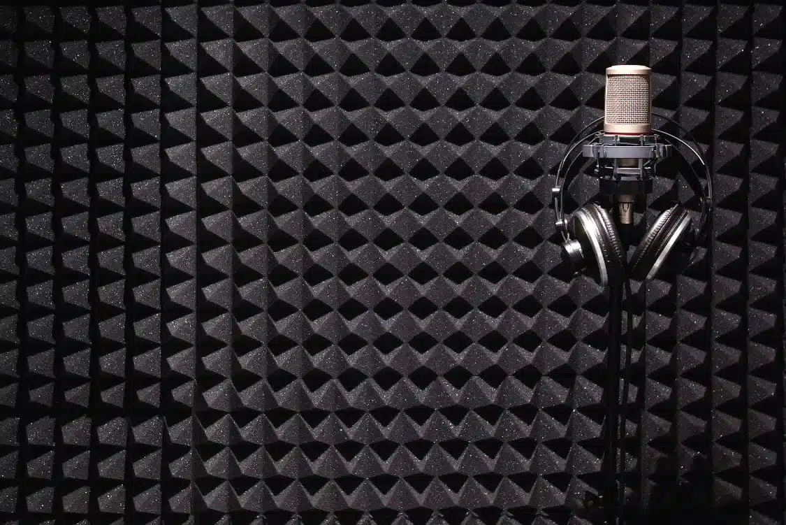 A microphone and headphones on a stand are placed in front of a wall covered in black acoustic foam panels in a soundproof room.
